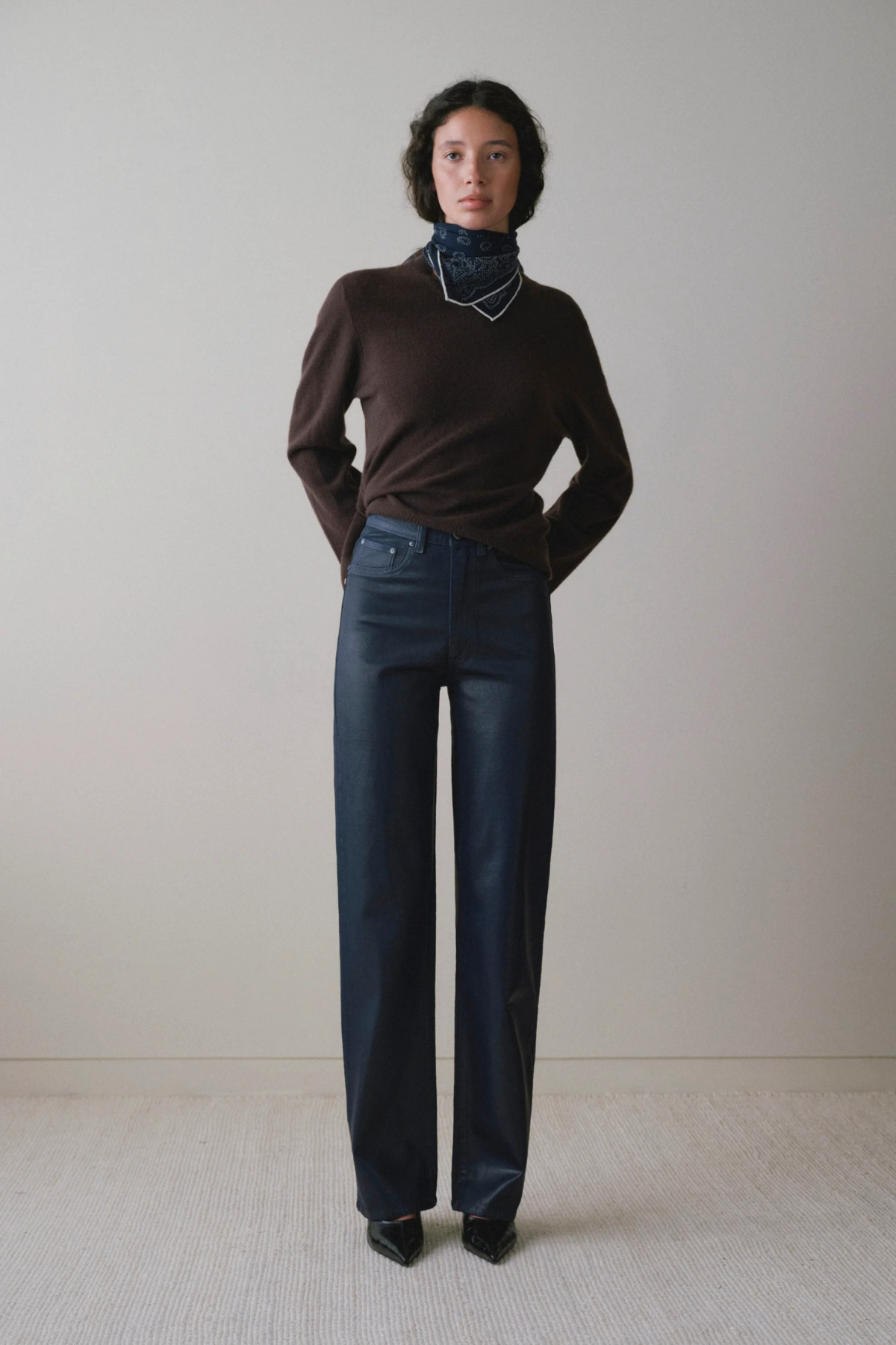A model stands against a plain light beige wall wearing a dark brown long-sleeve turtleneck sweater, dark blue wide-leg jeans, and black pointed shoes.