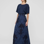 A woman modeling a navy blue two-piece outfit consisting of a short-sleeve top and a high-waisted maxi skirt with a floral jacquard pattern, paired with black strappy heels against a light gray studio background.