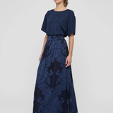 A woman modeling a navy blue two-piece outfit consisting of a short-sleeve top and a high-waisted maxi skirt with a floral jacquard pattern, paired with black strappy heels against a light gray studio background.