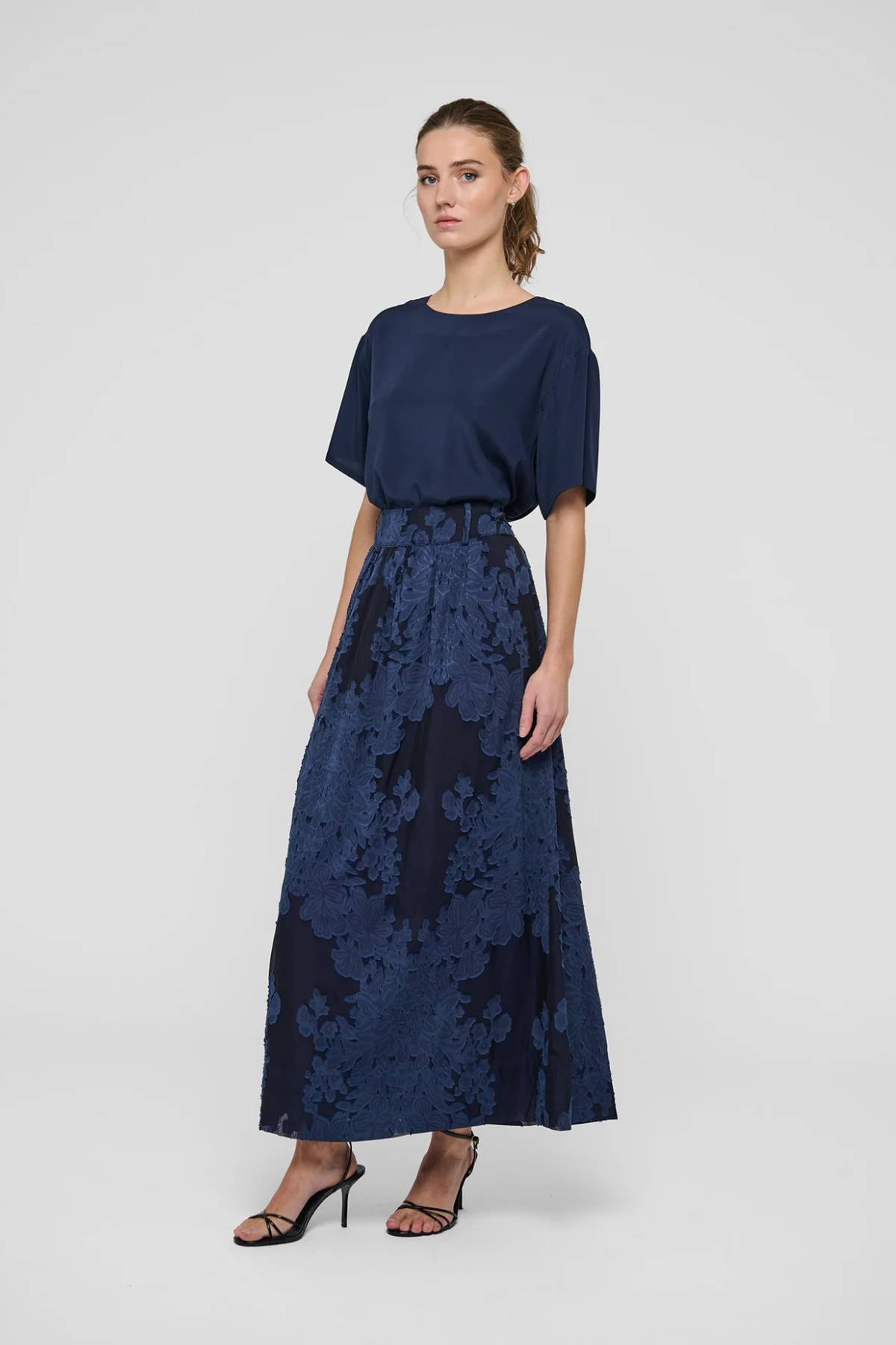 A woman modeling a navy blue two-piece outfit consisting of a short-sleeve top and a high-waisted maxi skirt with a floral jacquard pattern, paired with black strappy heels against a light gray studio background.