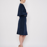A woman stands in profile wearing a dark navy velvet dress with a short cape-style yoke and elbow-length sleeves. The dress has a matching belt with gold-tone buttons at the waist.