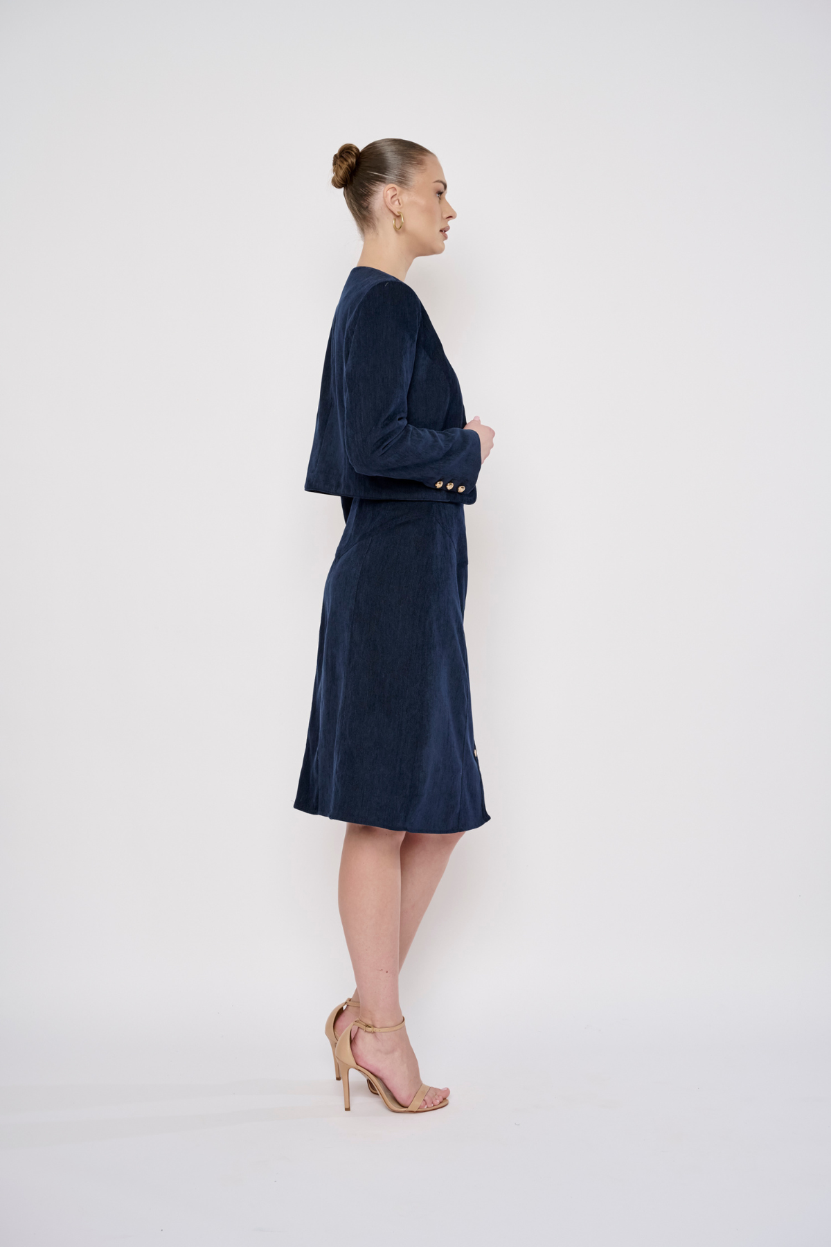 A woman stands in profile wearing a dark navy velvet dress with a short cape-style yoke and elbow-length sleeves. The dress has a matching belt with gold-tone buttons at the waist.