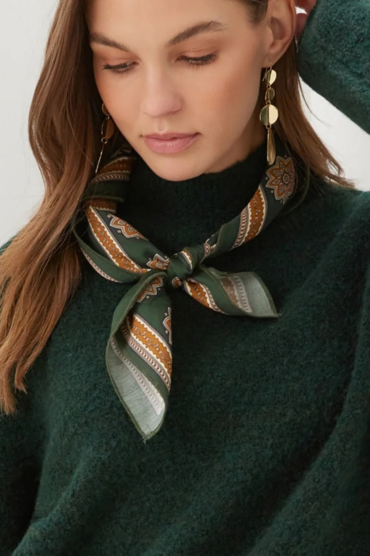 A close-up of a woman wearing a dark green knit sweater and a patterned silk scarf tied around her neck. The scarf features green, gold, and brown geometric and floral motifs.