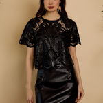 A woman modeling a black short-sleeve top with a sheer floral lace overlay and a black faux-leather mini skirt, standing against a warm beige background.