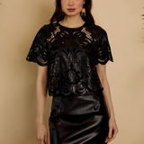 A woman modeling a black short-sleeve top with a sheer floral lace overlay and a black faux-leather mini skirt, standing against a warm beige background.