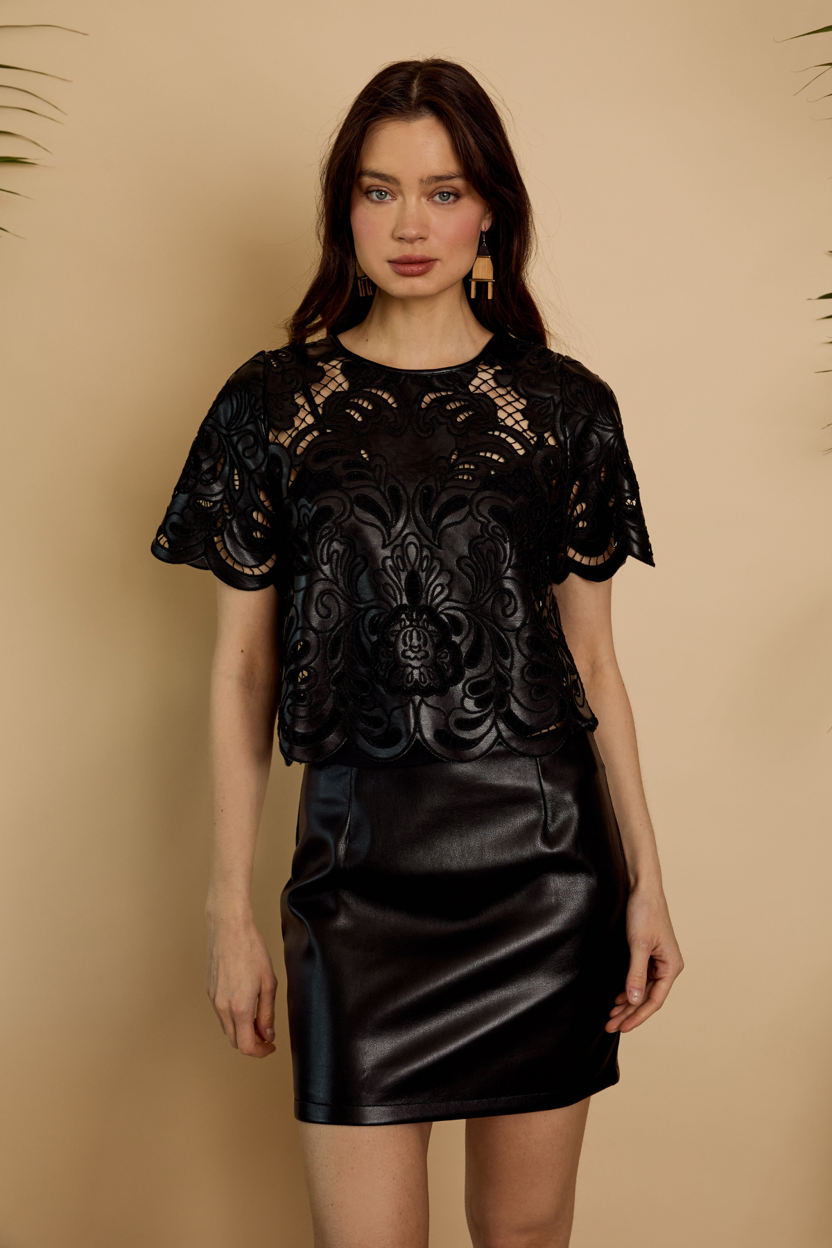A woman modeling a black short-sleeve top with a sheer floral lace overlay and a black faux-leather mini skirt, standing against a warm beige background.