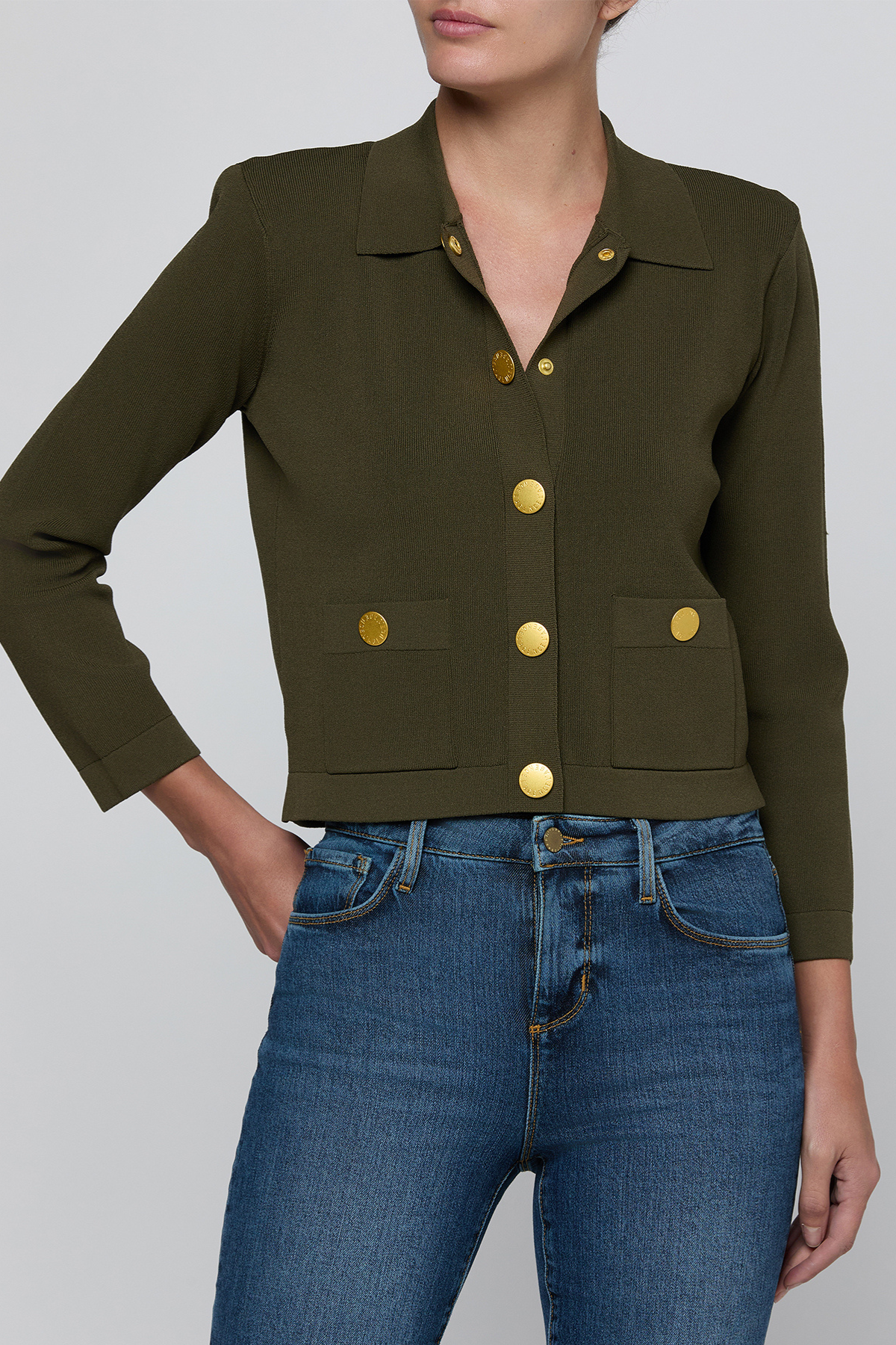 A woman wearing an olive green cropped jacket with gold-tone buttons and blue jeans, shown from the neck down against a light gray studio background.