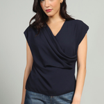A woman modeling a navy blue draped sleeveless top with blue jeans against a light gray studio background.