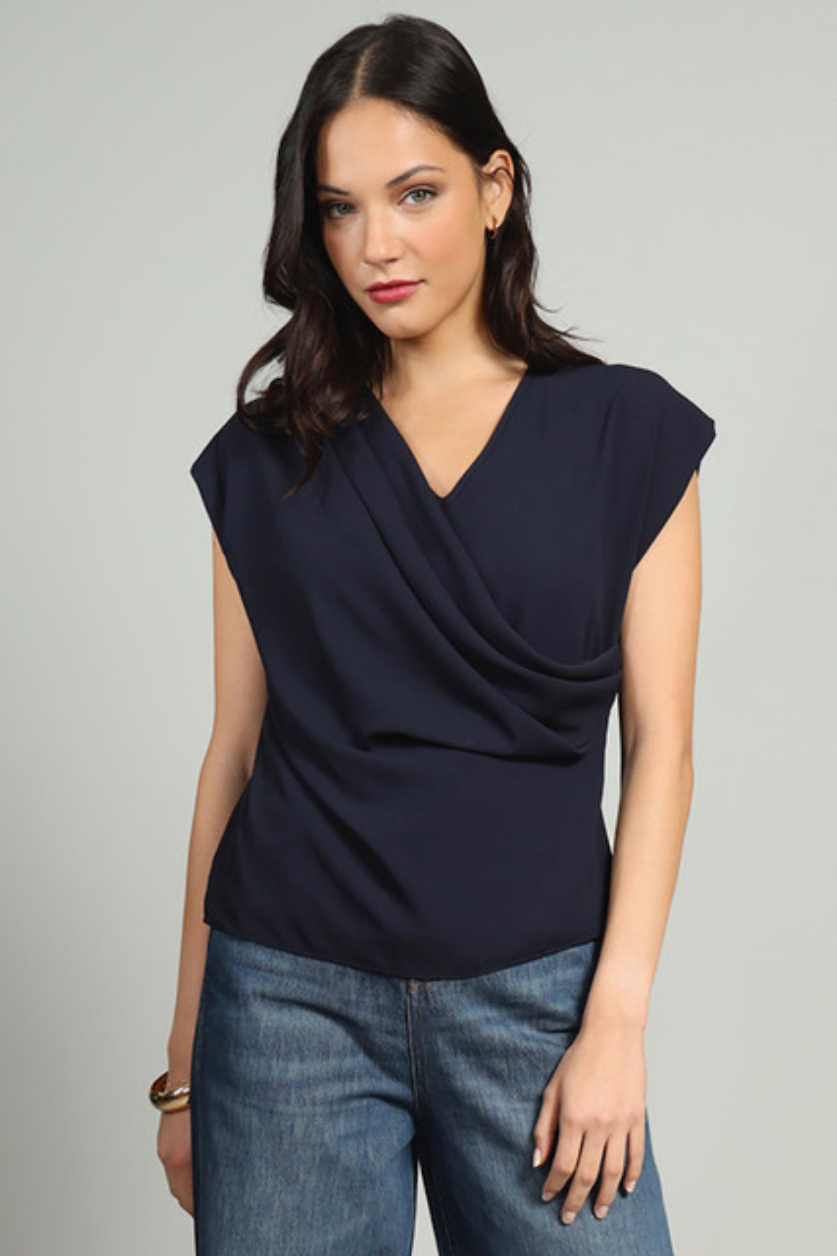 A woman modeling a navy blue draped sleeveless top with blue jeans against a light gray studio background.