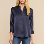 A blonde model wearing a dark navy blue satin button-down shirt with three-quarter sleeves and blue jeans, standing against a solid beige background.
