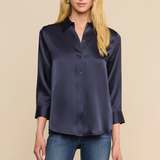 A blonde model wearing a dark navy blue satin button-down shirt with three-quarter sleeves and blue jeans, standing against a solid beige background.