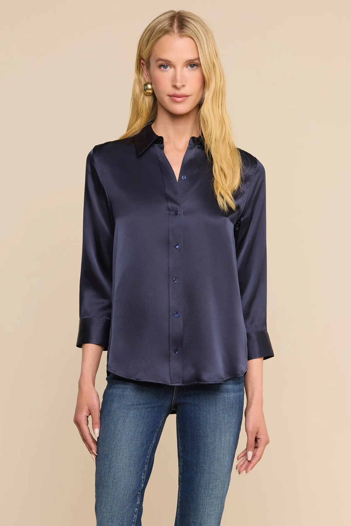 A blonde model wearing a dark navy blue satin button-down shirt with three-quarter sleeves and blue jeans, standing against a solid beige background.
