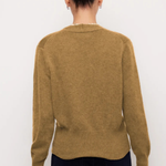 A woman viewed from the back wearing a camel brown knit sweater and dark navy pants, standing against a plain white background.