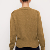 A woman viewed from the back wearing a camel brown knit sweater and dark navy pants, standing against a plain white background.