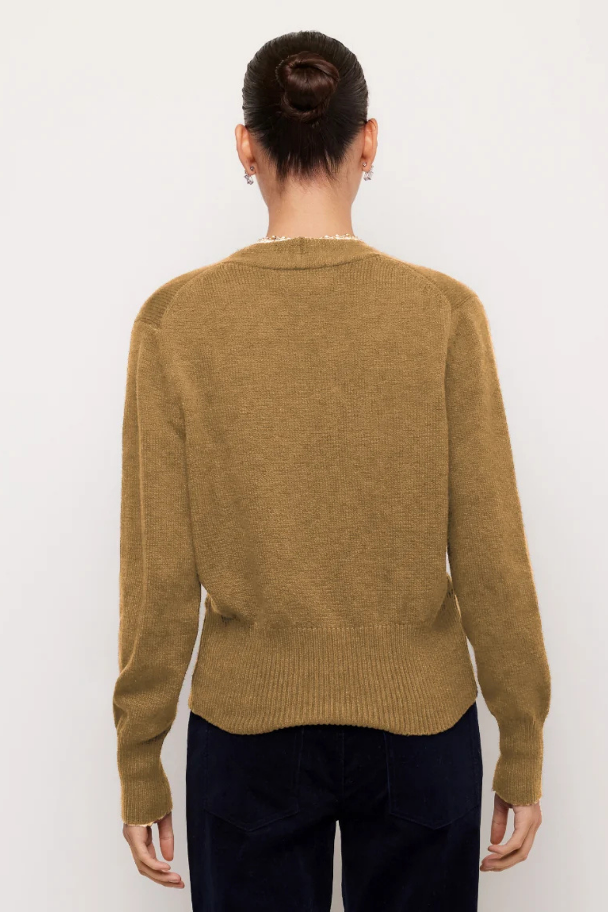 A woman viewed from the back wearing a camel brown knit sweater and dark navy pants, standing against a plain white background.
