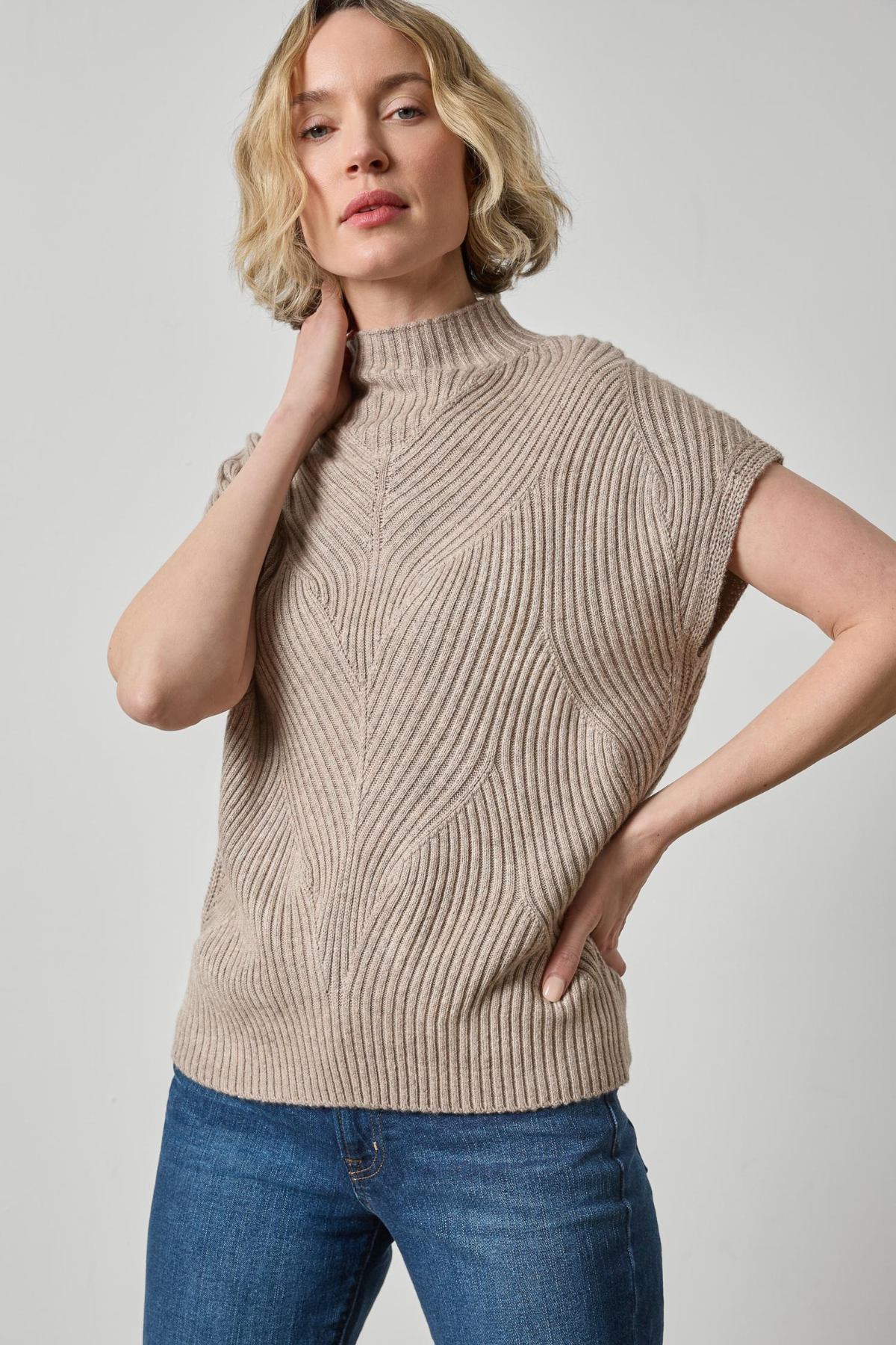 A woman wearing a beige ribbed knit mock-neck sweater vest with short sleeves, paired with blue jeans, against a light gray studio background.