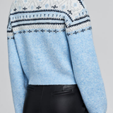 A woman wearing a light blue cropped knit sweater with a white and navy Fair Isle pattern across the shoulders and upper back, paired with black pants, shown from a rear three-quarter angle against a light gray studio background.