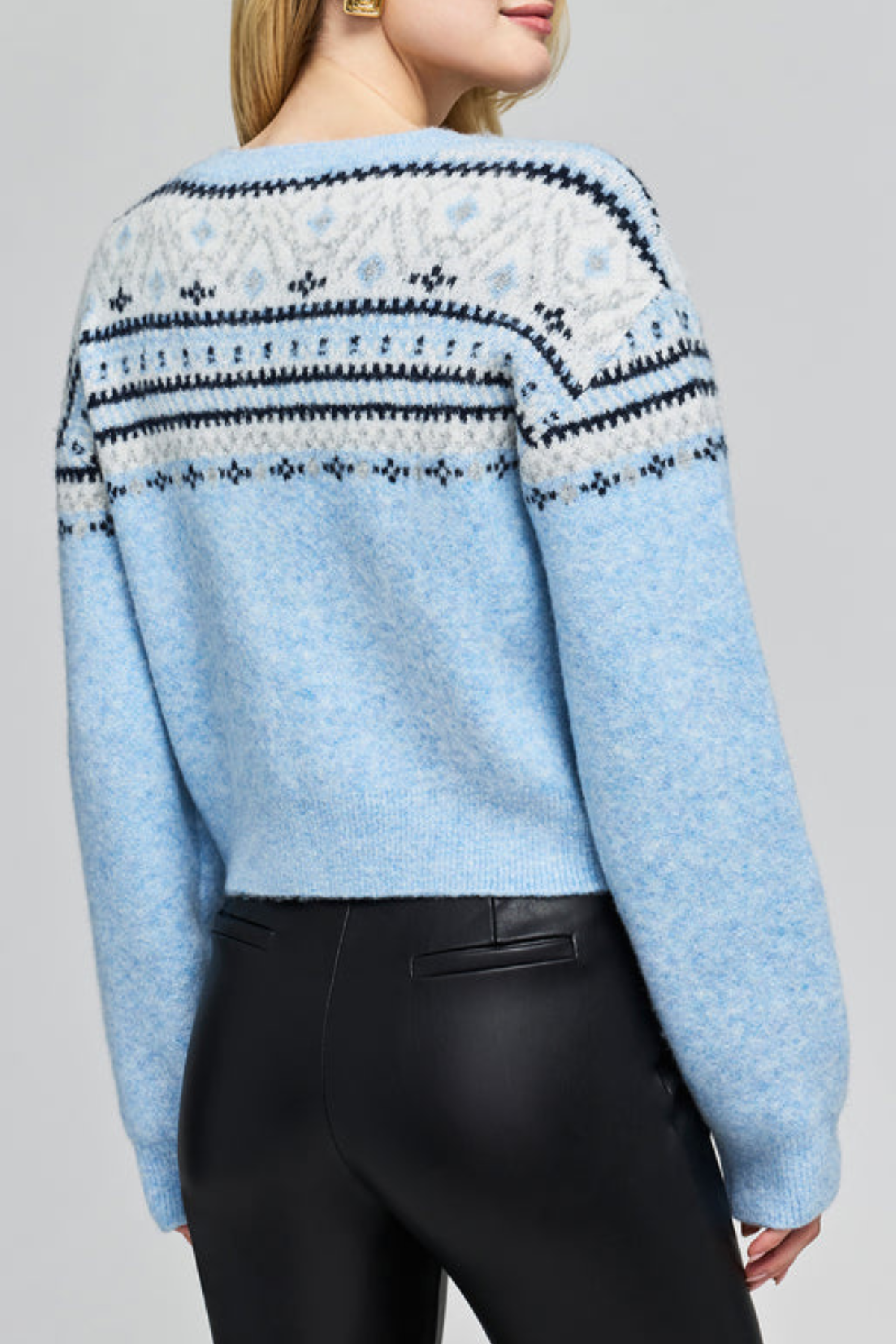 A woman wearing a light blue cropped knit sweater with a white and navy Fair Isle pattern across the shoulders and upper back, paired with black pants, shown from a rear three-quarter angle against a light gray studio background.