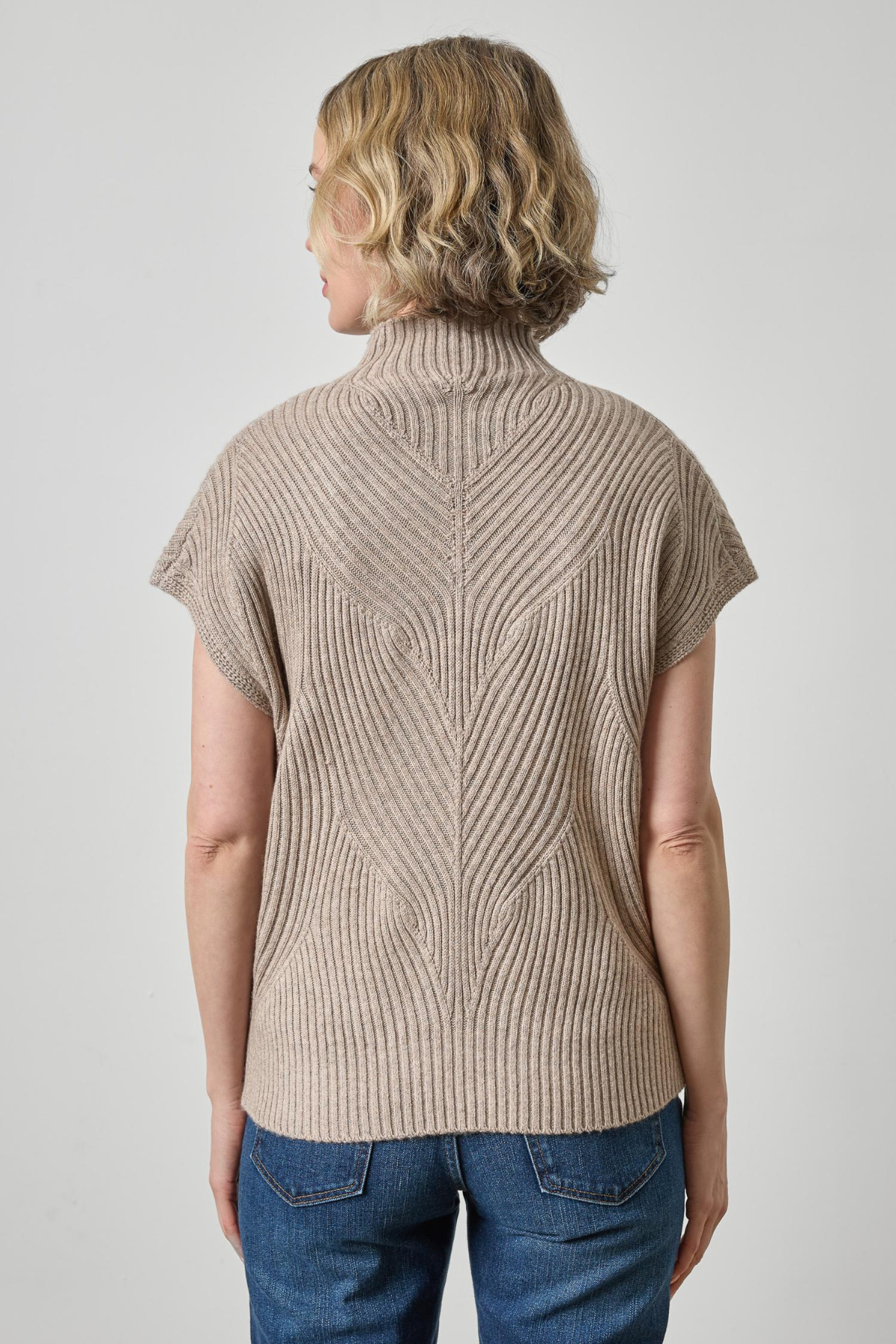 A woman shown from the back wearing a beige short-sleeve ribbed knit mock neck sweater and blue jeans against a light gray studio background.