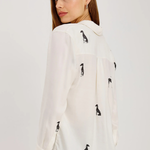A woman wearing a white long-sleeve blouse with black dog silhouettes printed all over it, shown from a side-back angle against a plain light gray studio background.