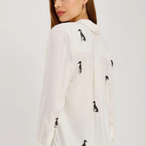 A woman wearing a white long-sleeve blouse with black dog silhouettes printed all over it, shown from a side-back angle against a plain light gray studio background.