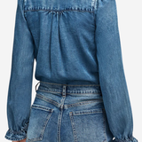 A close-up back view of a model wearing a blue denim long-sleeve blouse with gathered puff sleeves and matching blue denim jeans.