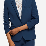 A woman wearing a tailored navy blue blazer with matching navy trousers and a white lace top, shown against a light gray studio background.
