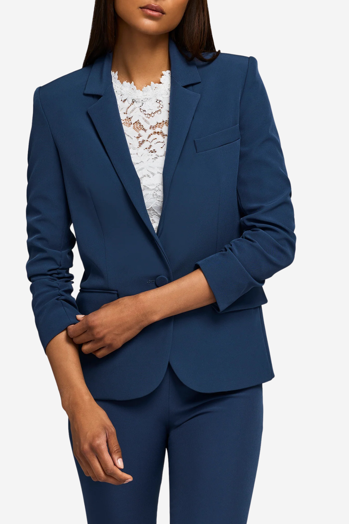 A woman wearing a tailored navy blue blazer with matching navy trousers and a white lace top, shown against a light gray studio background.