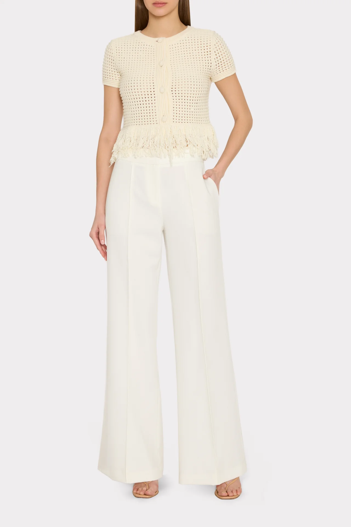 A model wearing a cream-colored short-sleeve knit top with a fringed peplum hem and matching cream wide-leg trousers, shown against a light gray studio background.