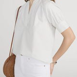 Woman wearing a white blouse and white pants with a brown woven bag on a plain background