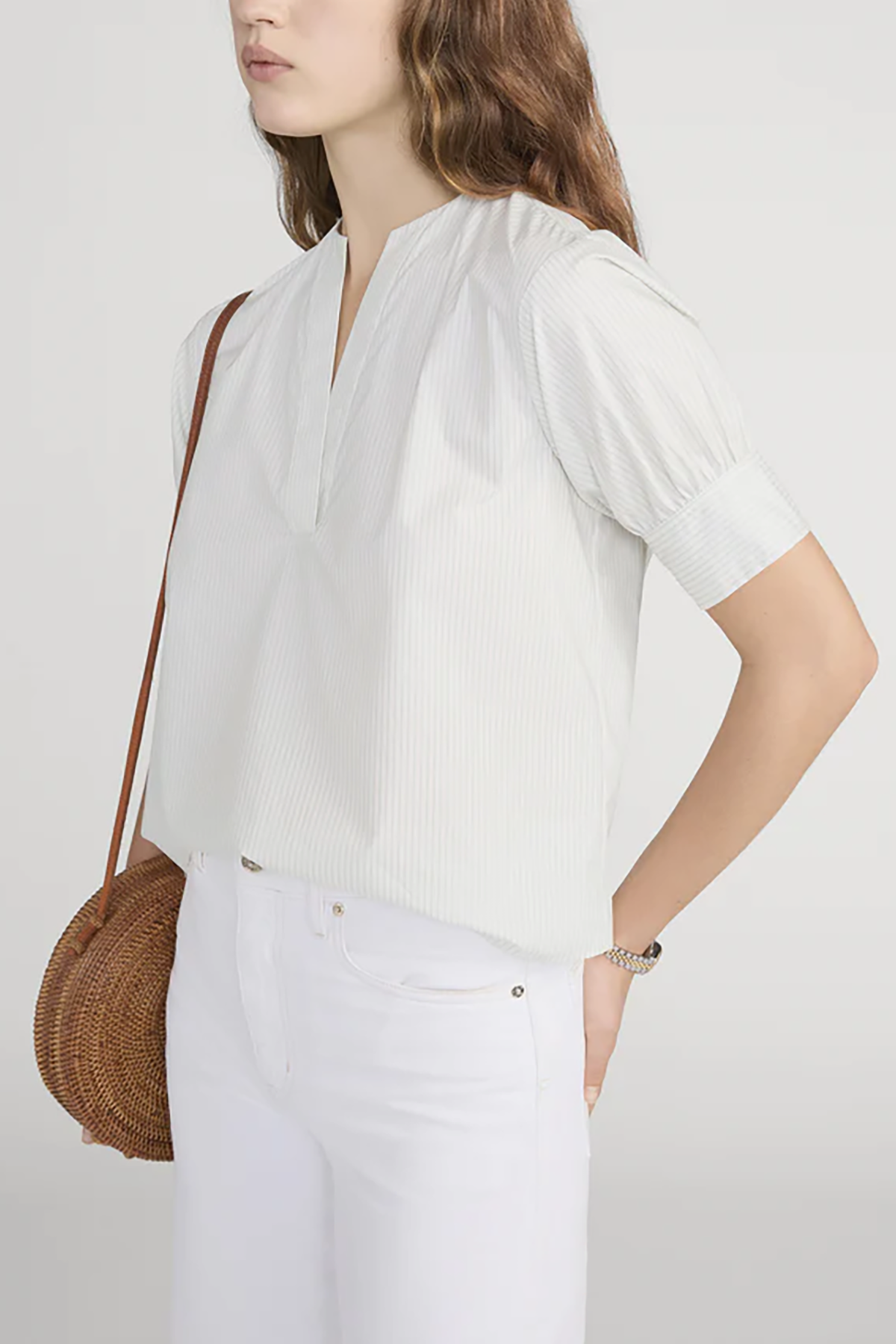 Woman wearing a white blouse and white pants with a brown woven bag on a plain background