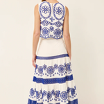 A woman viewed from the back wearing a sleeveless white maxi dress with intricate blue floral embroidery and tiered ruffles, paired with brown sandals, against a plain off-white studio background.