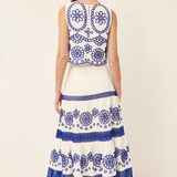 A woman viewed from the back wearing a sleeveless white maxi dress with intricate blue floral embroidery and tiered ruffles, paired with brown sandals, against a plain off-white studio background.