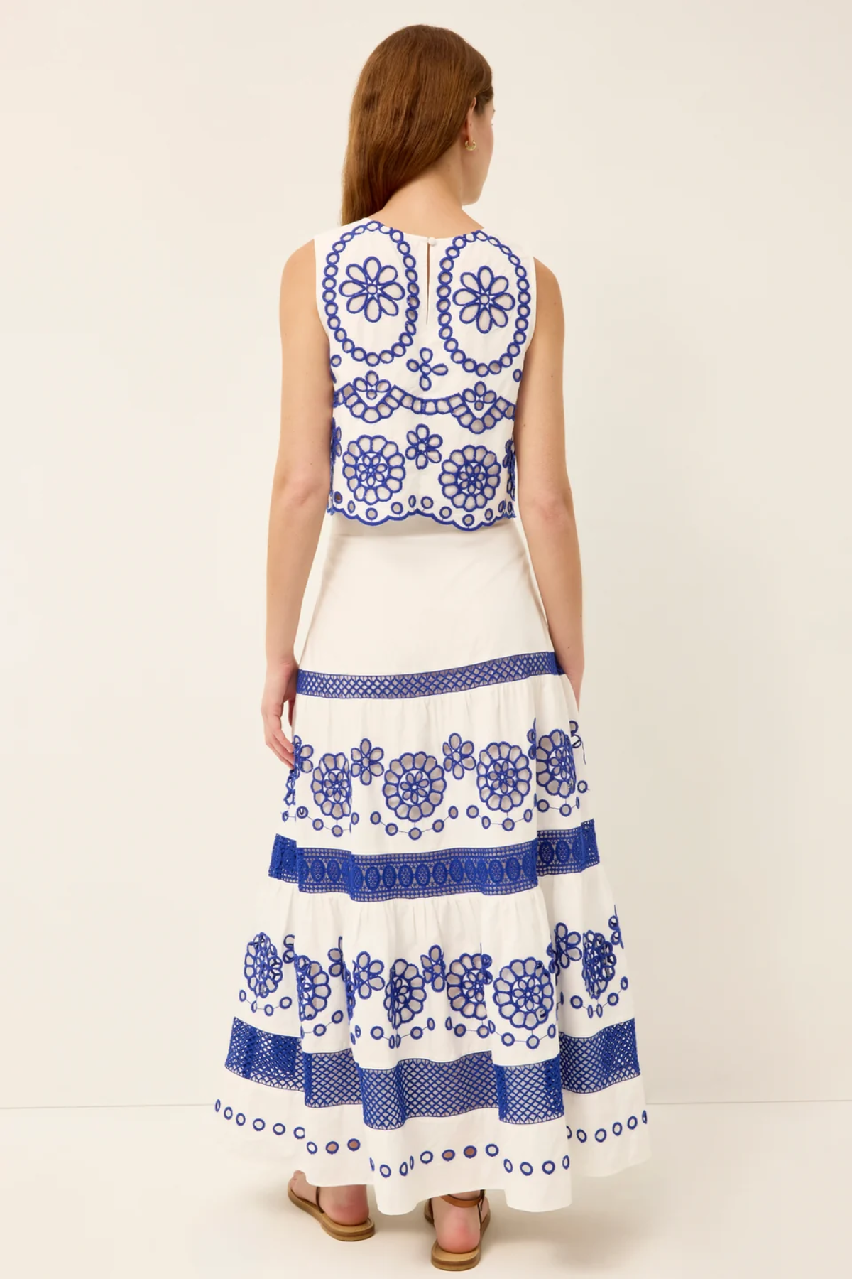 A woman viewed from the back wearing a sleeveless white maxi dress with intricate blue floral embroidery and tiered ruffles, paired with brown sandals, against a plain off-white studio background.