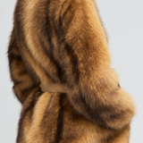A close-up back view of a model wearing a luxurious brown and tan fur jacket with a matching belt, set against a plain light gray background.