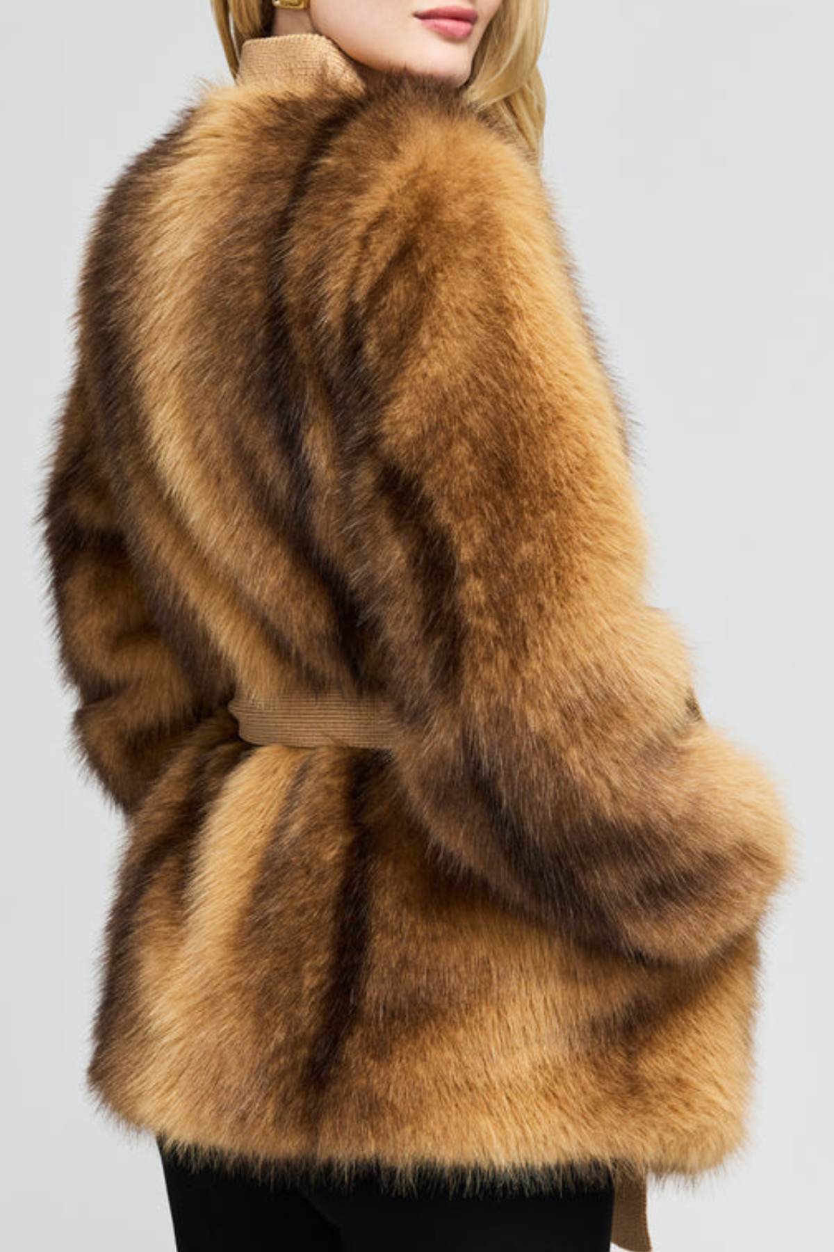 A close-up back view of a model wearing a luxurious brown and tan fur jacket with a matching belt, set against a plain light gray background.