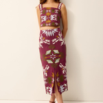 A woman modeling a matching two-piece set featuring a sleeveless crop top and a midi skirt, both in a deep magenta color with a white floral and green botanical pattern, set against a plain off-white studio background.