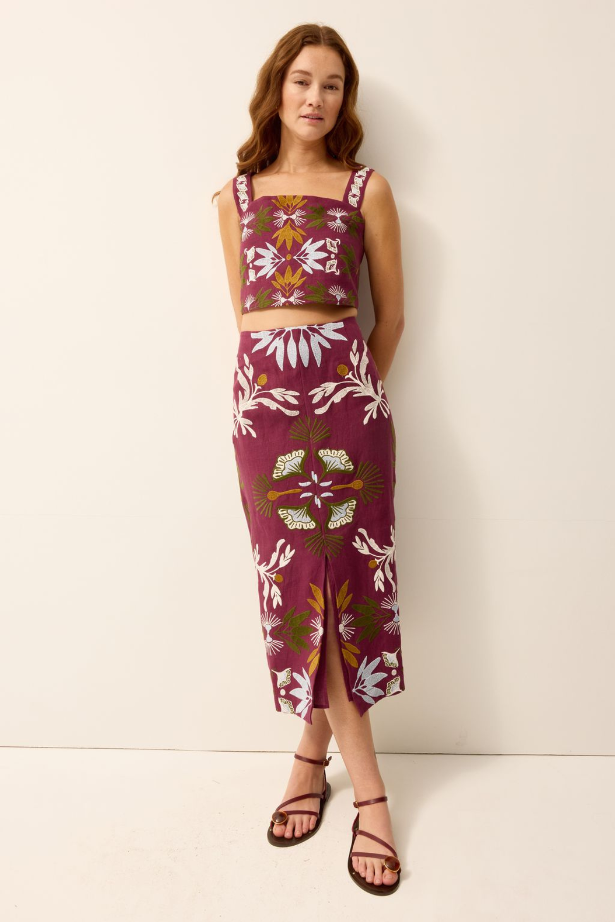 A woman modeling a matching two-piece set featuring a sleeveless crop top and a midi skirt, both in a deep magenta color with a white floral and green botanical pattern, set against a plain off-white studio background.