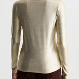 A woman shown from the back wearing a metallic champagne-gold turtleneck sweater and dark brown pants against a plain light gray studio background.