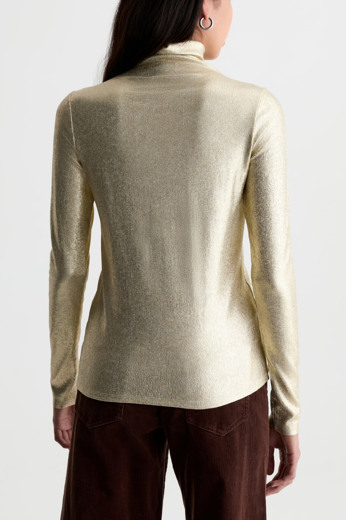 A woman shown from the back wearing a metallic champagne-gold turtleneck sweater and dark brown pants against a plain light gray studio background.