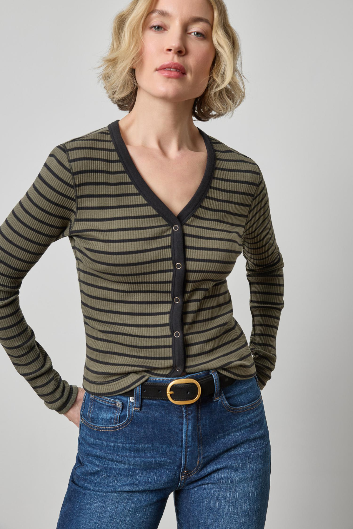 A woman wearing a ribbed long-sleeve cardigan with horizontal olive green and black stripes, paired with blue jeans and a black belt, against a plain light gray studio background.