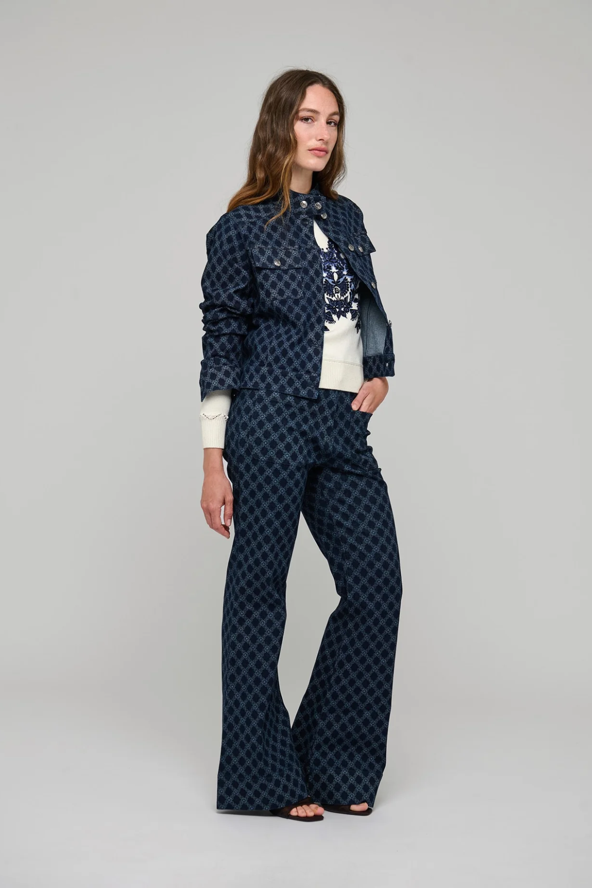 A woman modeling a matching navy blue patterned jacket and wide-leg trousers set, styled with a white top, against a neutral gray studio background.