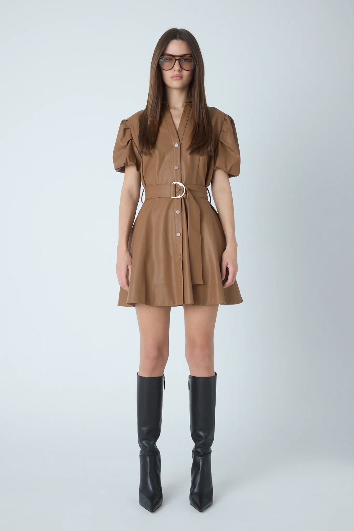 A woman modeling a brown mini shirt dress with puff sleeves, a button front, and a matching belt with a silver buckle, paired with black knee-high boots against a light gray studio background.