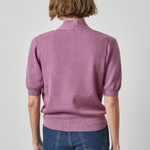 A model viewed from the back wearing a dusty purple short-sleeve knit sweater with a mock neck and blue jeans, set against a plain light gray studio background.