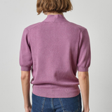 A model viewed from the back wearing a dusty purple short-sleeve knit sweater with a mock neck and blue jeans, set against a plain light gray studio background.