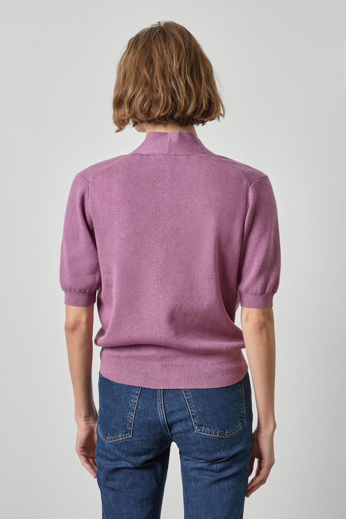 A model viewed from the back wearing a dusty purple short-sleeve knit sweater with a mock neck and blue jeans, set against a plain light gray studio background.