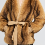A woman wearing a luxurious camel-brown faux fur wrap coat with a matching beige knit tie belt, styled with black pants against a light gray background.
