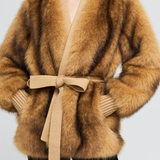 A woman wearing a luxurious camel-brown faux fur wrap coat with a matching beige knit tie belt, styled with black pants against a light gray background.