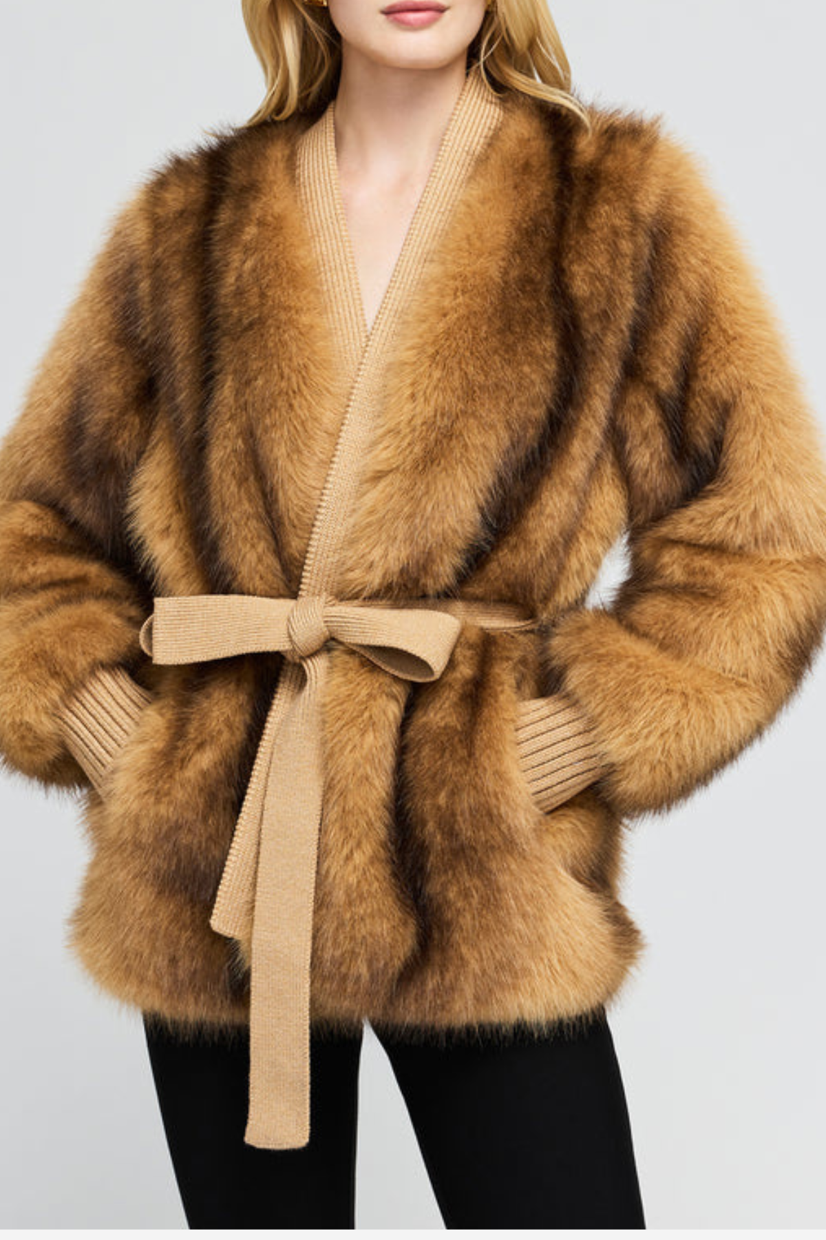 A woman wearing a luxurious camel-brown faux fur wrap coat with a matching beige knit tie belt, styled with black pants against a light gray background.