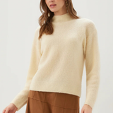 A woman wearing a cream-colored, fuzzy mock-neck knit sweater and brown wide-leg trousers, photographed against a clean, light gray studio background.